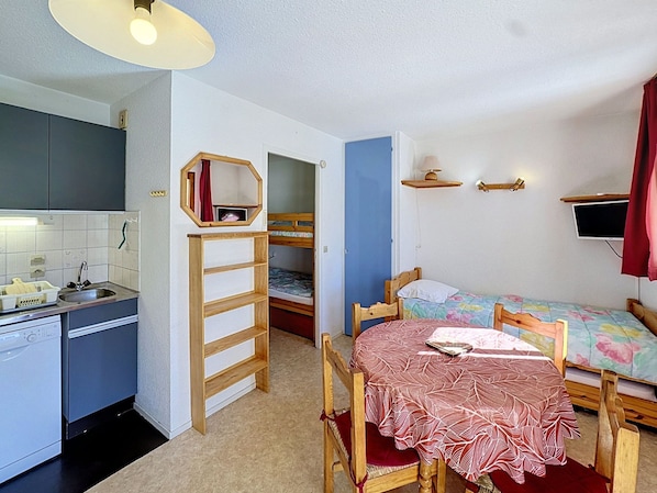 Une pièce compacte comprenant un coin cuisine, un lit superposé, un lit simple, une table recouverte d’une nappe à motifs et un téléviseur fixé au mur.