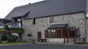 A stone building with a sloped roof, a balcony, and a small garden area with flowers and shrubs.