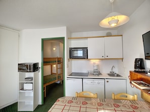 Cocina americana compacta con microondas, cafetera y lavamanos. Se ve una litera a través de una puerta abierta.