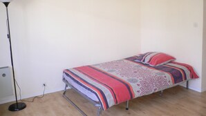 Una habitación pequeña con una cama individual de estructura metálica, una lámpara de pie y un enchufe en la pared.