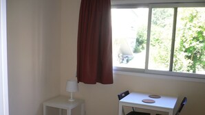 Una habitación pequeña con una mesa blanca, dos sillas negras, una lámpara blanca y una ventana con cortina roja.