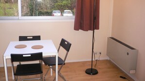 Una habitación con una mesa blanca y dos sillas negras, una lámpara de pie, una ventana con vista a coches y árboles, y un radiador en la pared.