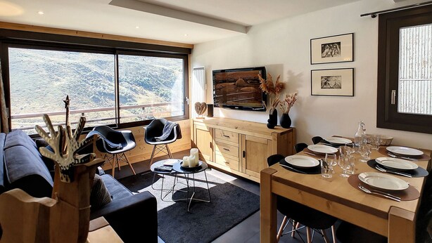 Een moderne woonkamer met een groot raam, een eethoek met een houten tafel gedekt voor vier personen, en een gezellige zithoek met een bank en fauteuils.