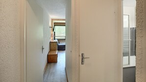 Un estrecho pasillo que lleva a un dormitorio con ventana, un banco de madera y una puerta con pomo metálico.