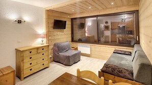 Una habitación acogedora con techo de madera, un ventanal con persianas, un sofá, una silla pequeña y un aparador de madera con tiradores con forma de corazón.