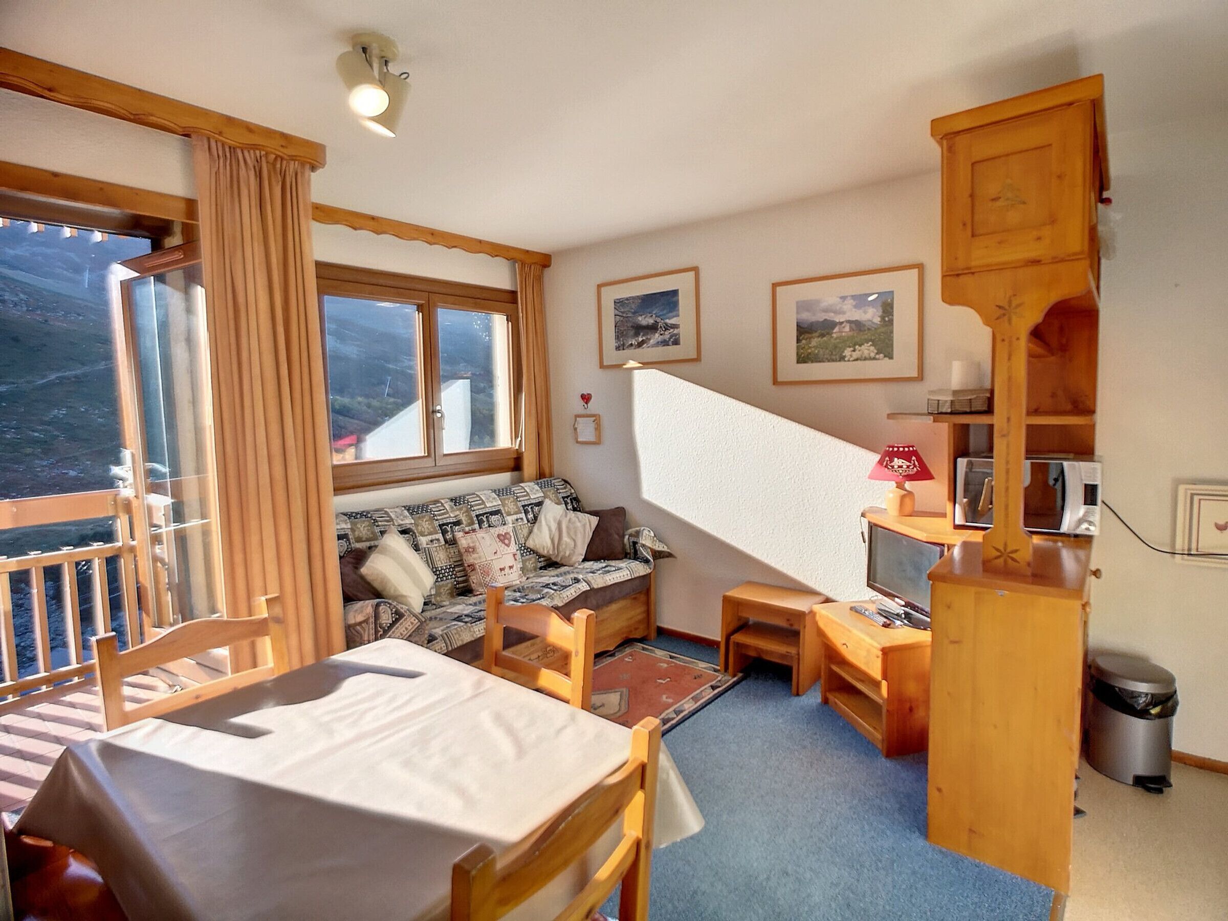 A cozy room with a dining table, a sofa, a small desk, and a trash bin. There are windows with curtains, a lamp, and framed pictures on the wall.