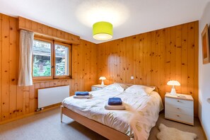 A wooden-paneled bedroom with a bed, two bedside tables with lamps, a window with curtains, and a radiator. - Charming flat located in the quiet region of Chemin des Vernes, in 5 minutes of the ski lift of Médr (Verbier)