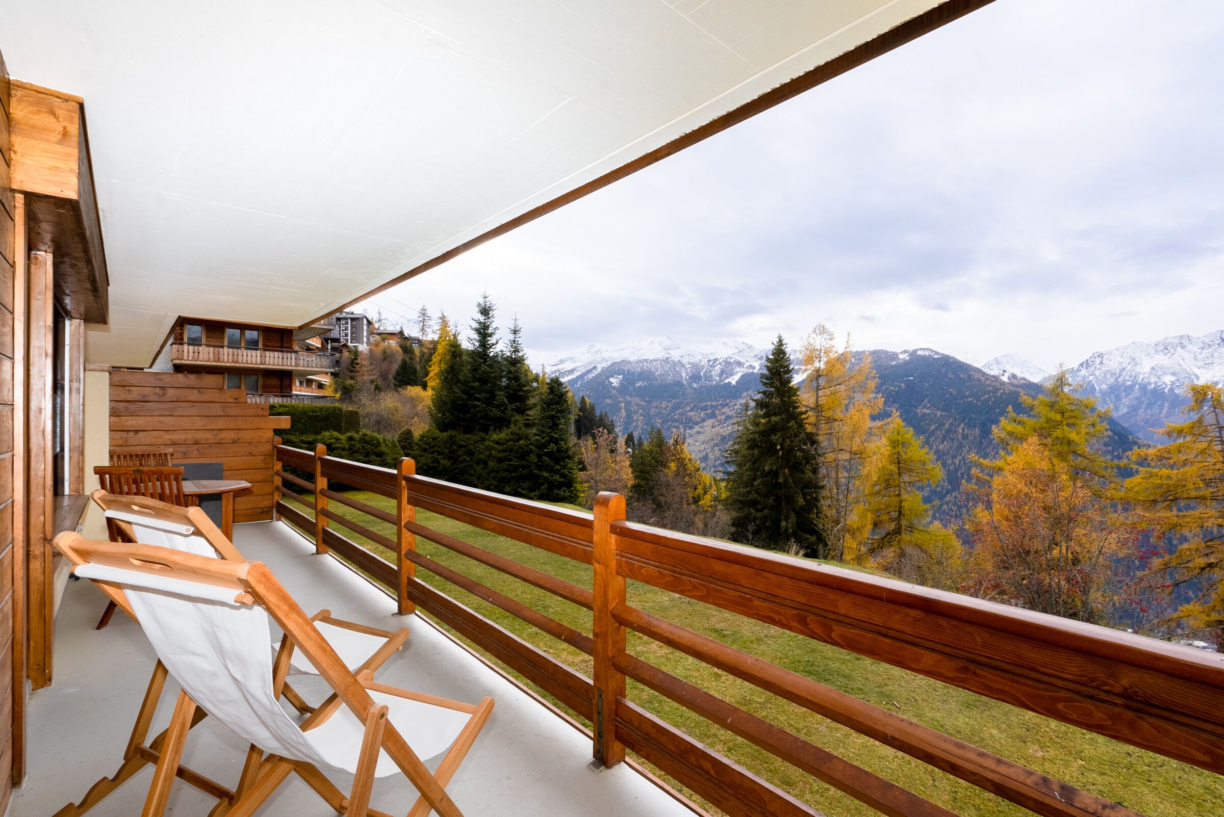 Una terraza de madera con asientos, con vista a un paisaje montañoso con follaje otoñal y picos nevados.