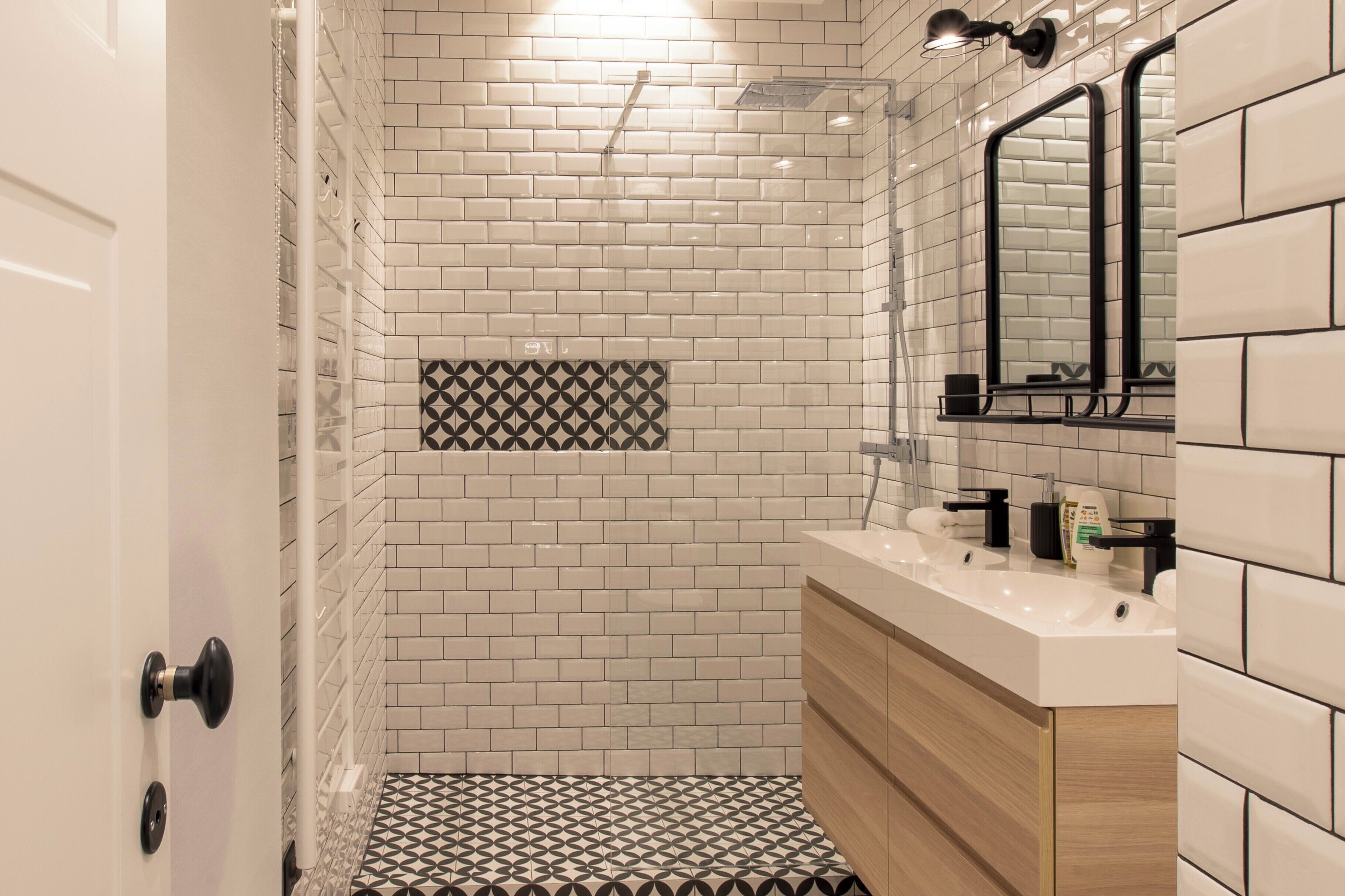 Un baño moderno con azulejos blancos tipo metro, un detalle de azulejos con diseño geométrico, un lavamanos blanco con accesorios negros y un espejo con marco negro.