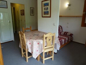 A dining area with a table covered by a floral tablecloth, surrounded by wooden chairs. There is a sofa with a patterned cover in the background.
