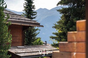 A wooden building with a sloped roof, surrounded by evergreen trees and mountains in the background.