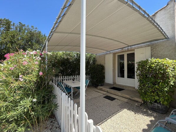A covered patio area with a table and chairs, surrounded by a white picket fence and various plants.