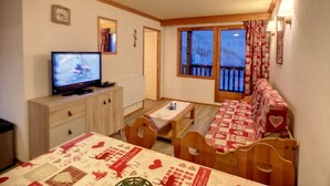 Una habitación acogedora con televisor, muebles de madera, una mesa con mantel estampado con corazones y un balcón con cortinas.