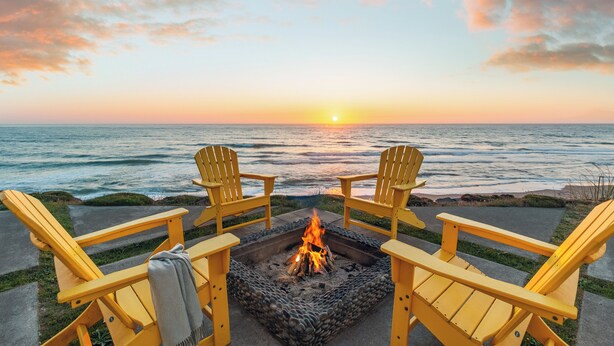 A sunset view from a deck with two yellow Adirondack chairs facing a fire pit. - Ocean Front Luxury Home (Gleneden Beach)