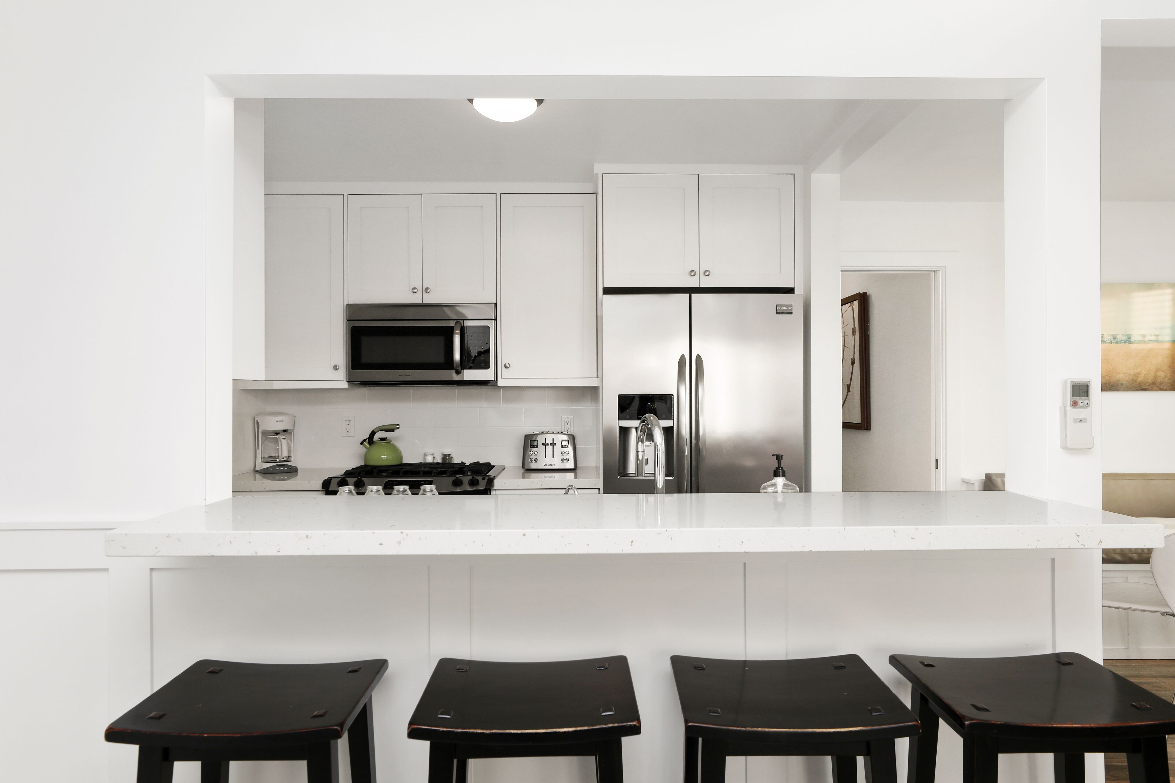 A modern kitchen with white cabinetry, a stainless steel refrigerator, a microwave, and a coffee maker. There are four wooden stools at the island.