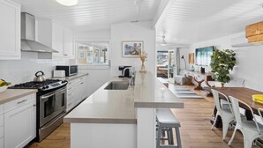 A modern kitchen with white cabinetry, a central island, stainless steel appliances, and a dining area with a wooden table and chairs. - 37th Street beach house, steps to sand - Air conditioning, bikes and parking! (Newport Beach)