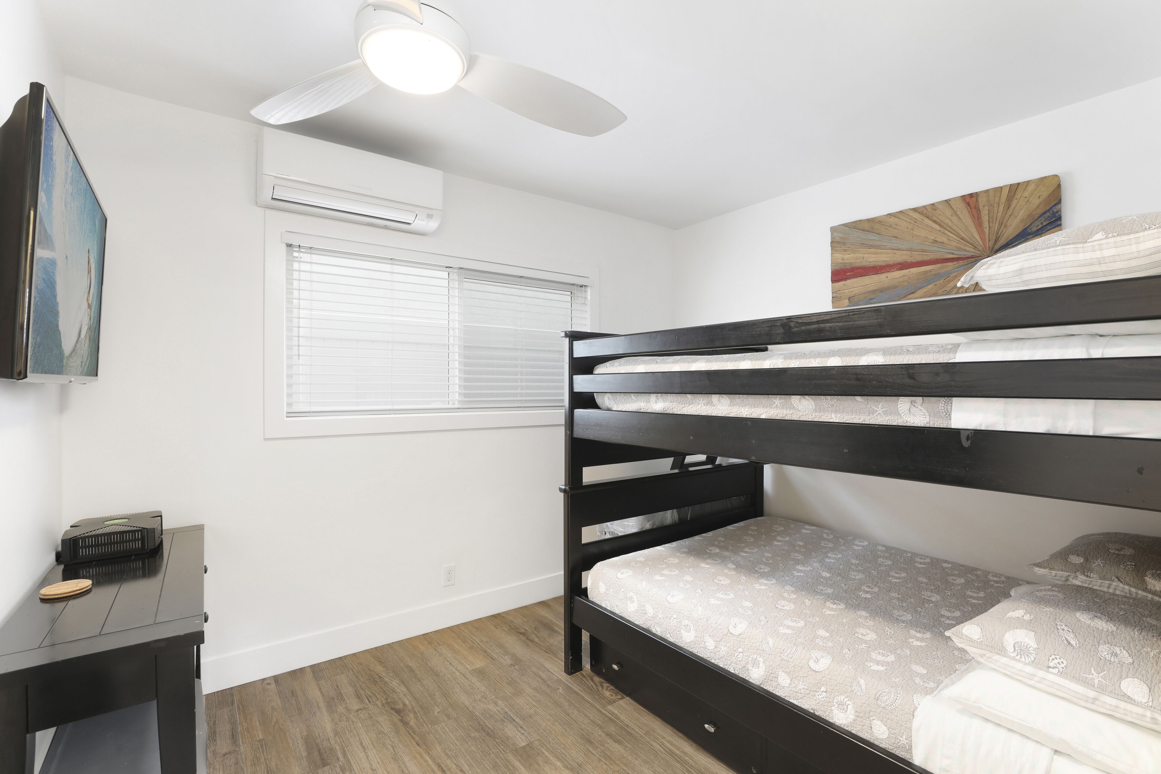 Uma cama de beliche com ventilador de teto, janela com persianas, uma mesa pequena com rádio e um quadro fixado na parede.
