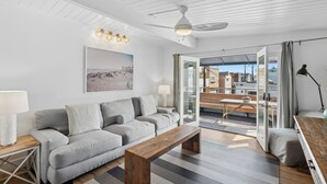 A modern living room with a grey sofa, a wooden coffee table, and a ceiling fan. There is a large window with a view of buildings outside. - 37th Street beach house, steps to sand - Air conditioning, bikes and parking! (Newport Beach)