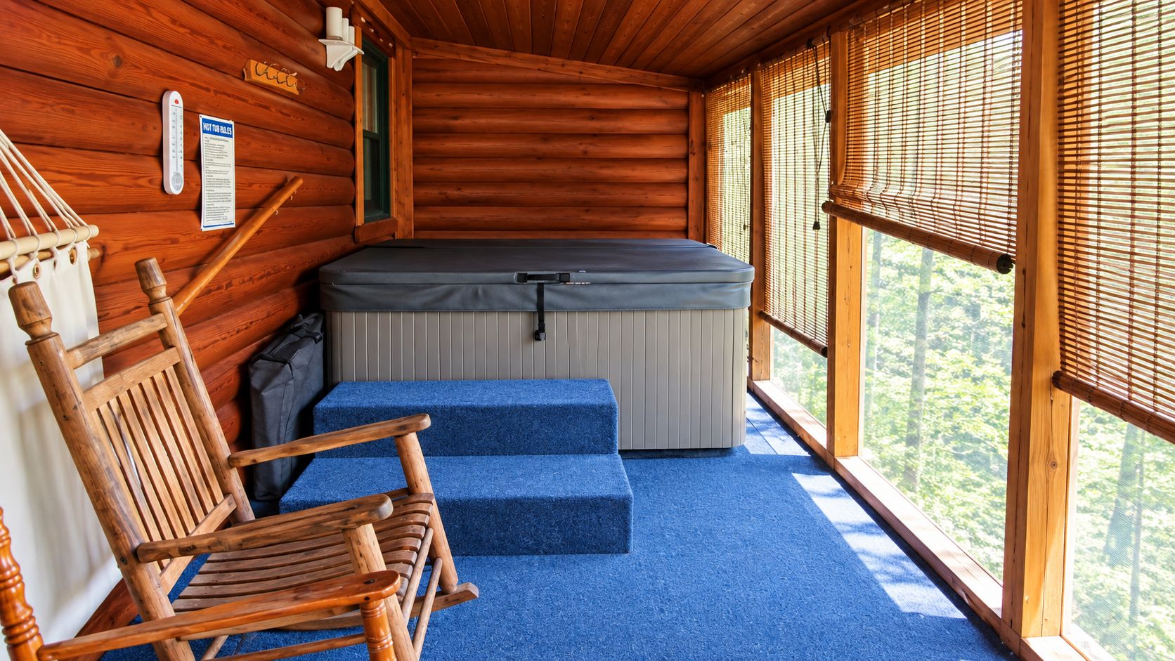 A log cabin interior with a hot tub, a wooden rocking chair, and a view of the forest through the windows.