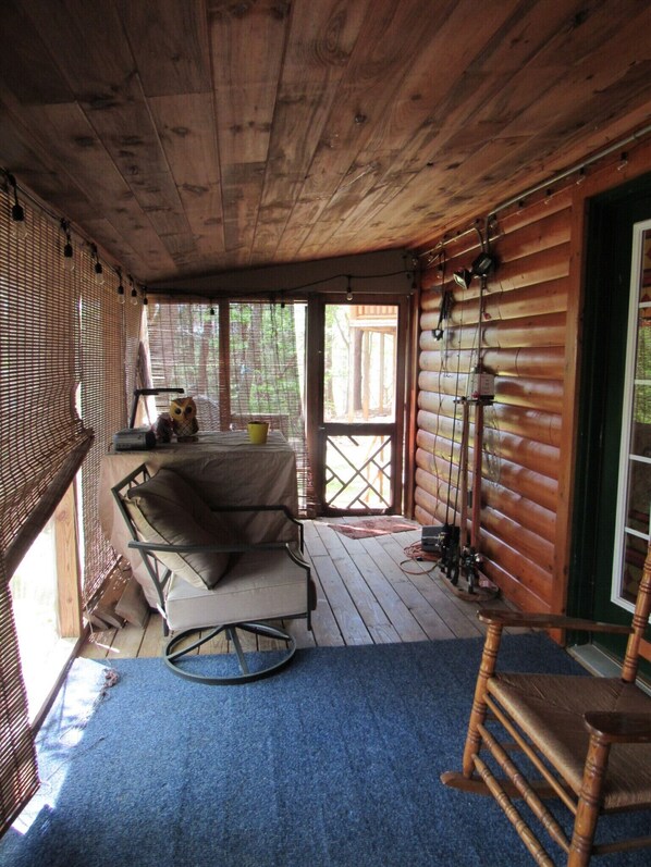 A wooden cabin with a covered porch, a cushioned chair, a small table, and a blue carpet.
