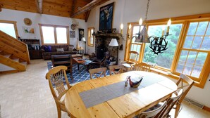 Ein rustikales Blockhaus-Interieur mit einem Holzespeisetisch, Stühlen, einem Kamin und einem großen Fenster mit Blick auf Grünflächen.