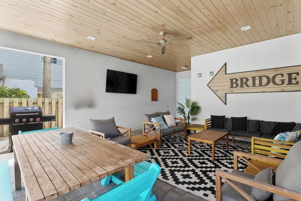 A modern outdoor living area with a wooden table, chairs, a sofa, and a black and white patterned rug. A television is mounted on the wall, and a sign with the word "BRIDGE" is visible.