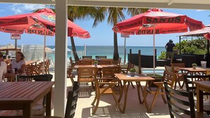 An outdoor seating area with wooden tables and chairs, red umbrellas, and a view of the ocean. - Beach Front Studio For Rent in Freeport Grand Bahamas , Port Lucaya (Freeport)