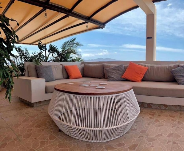 Ein Außenbereich mit Sitzgelegenheit, einem runden Gewebetisch, einer gepolsterten Couch und einer Pergola darüber.