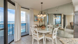 A dining area with a round table, white chairs, and a view of the ocean through large windows. - Come Enjoy the BEACH! One Bedroom ~ Phoenix VII (Orange Beach)