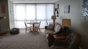 A living room with a sofa, a coffee table, and a dining table with chairs. There is a window with blinds, a lamp, and wall art. - Closest building to the main lift. (Frisco)