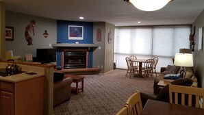 A living room with a fireplace, a dining area with chairs, a kitchen area with a kettle, and a large window with blinds. - Closest building to the main lift. (Frisco)