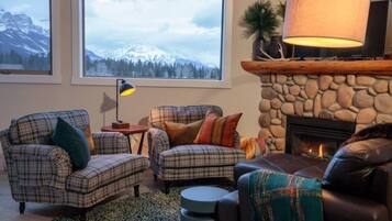 A cozy living room with a fireplace, two plaid armchairs, a coffee table, and a view of snow-capped mountains through large windows.