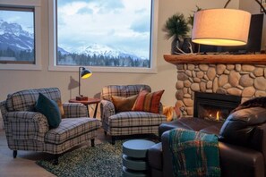Una sala de estar acogedora con chimenea, dos sillones a cuadros, una mesita de centro y vistas a montañas nevadas a través de amplios ventanales.
