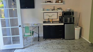 Une cuisine compacte équipée d’un four à micro-ondes, d’un grille-pain et d’un réfrigérateur. Une chaise est placée près de la porte, et une étagère au-dessus du comptoir contient divers ustensiles de cuisine.