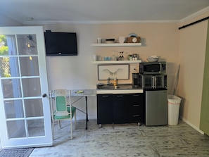 Een kleine keuken met een magnetron, broodrooster en koelkast. Een stoel staat bij de deur, en een plank boven het aanrecht bevat verschillende keukenbenodigdheden.