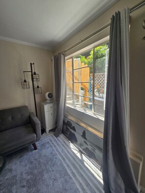 A room with a grey sofa, a small white cabinet, a floor lamp, and a large window with curtains.