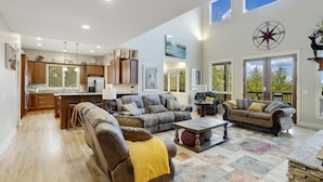 A spacious living room with a large brown leather sofa, a coffee table, and a fireplace. There is a kitchen area with wooden cabinets and a large window. - Large Luxury Home with Great Views (Banner Elk)