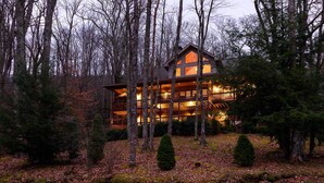 A multi-story log cabin with a warm-lit interior, surrounded by a forested area with fallen leaves on the ground. - Large Luxury Home with Great Views (Banner Elk)