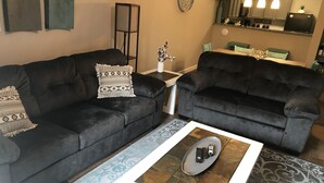 Ein Wohnzimmer mit zwei dunklen Sofas, einem Couchtisch mit Fernbedienungen und einer Küche im Hintergrund.