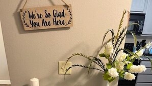 Ein Holzkonsolentisch mit einer weißen Vase, einer Kerze und einem Buch. Ein Schild darüber zeigt den Text 'We're So Glad You Are Here'.