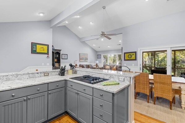 A modern kitchen with grey cabinets, a large island, and a dining area with wicker furniture. - Fairway Dunes 27 - Screen Porch Golf Course View! (Isle of Palms)