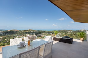 Eine Terrasse mit Esstisch aus Glas, weißen Korbstühlen, einer gepolsterten Sitzecke und Blick auf eine Küstenlandschaft.