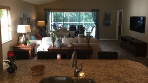 A spacious living room with a large window, a ceiling fan, and a flat-screen TV mounted on the wall. - 5⭐️ LUXURY BEACH VILLA with SECLUDED HEATED POOL+HOT TUB SURROUNDED by PALM TREES (Naples)
