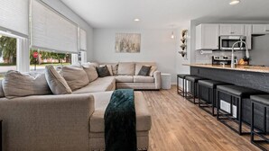 A modern living room with a sectional sofa, a kitchen area with bar stools, and a large window with blinds. - Walk-to-Lambeau Tailgate House (Green Bay)