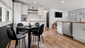 A modern kitchen with white cabinets, a dining table with chairs, a built-in oven, and a TV mounted on the wall. - Walk-to-Lambeau Tailgate House (Green Bay)