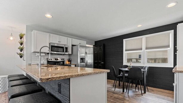 A modern kitchen with white cabinets, a granite countertop, stainless steel appliances, and a dining area with black chairs. - Walk-to-Lambeau Tailgate House (Green Bay)