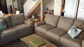 A living room with two beige sofas, a glass coffee table, and a staircase in the background.