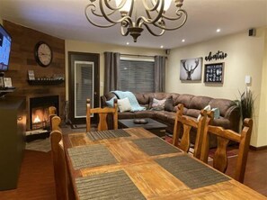 A living room with a fireplace, a large sectional sofa, a dining table with chairs, and a chandelier.