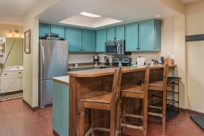 A compact kitchen with teal cabinets, a stainless steel refrigerator, a microwave, and a wooden bar area with chairs. - Giant Steps 63 - Walkout to the slopes (Brian Head)