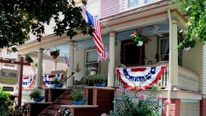 A two-story house with a porch decorated with American flags and a red, white, and blue banner. There are potted plants and flowers on the porch and steps. - Stay in the Heart of Cape May at the Charming Rose Suite (Cape May)